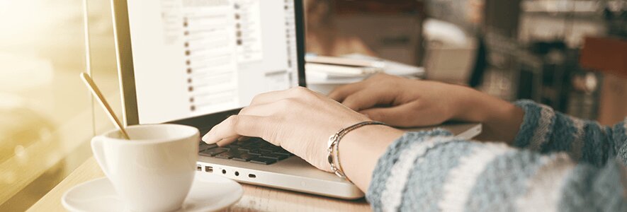 Eine Frau in einem gemütlichen Pullover tippt auf ihrem Laptop in einem Café, während neben ihr eine Tasse Kaffee mit Löffel steht.