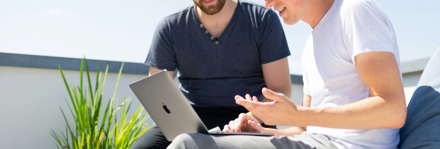 Zwei Männer sitzen draußen auf einer Terrasse mit einem MacBook auf dem Schoß und diskutieren angeregt.