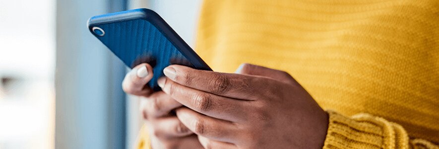 Eine Frau in einem gelben Strickpullover hält ein blaues Smartphone mit beiden Händen, während sie es betrachtet.