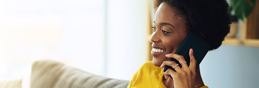 Eine Frau mit lockigem Haar und gelbem Oberteil hält ein Smartphone ans Ohr und lächelt. Sie sitzt entspannt auf einem Sofa in einem hellen Wohnzimmer.