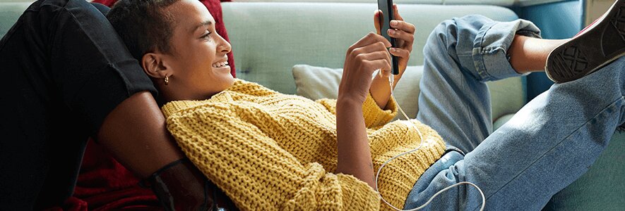 Eine junge Frau in einem gelben Strickpullover liegt entspannt auf einem Sofa, hält ihr Smartphone in der Hand und hört Musik mit weißen Kopfhörern.