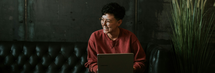 Eine Person mit Brille und rotem Pullover sitzt auf einem dunklen Ledersofa, hält einen Laptop auf dem Schoß und lacht herzlich in einem stilvollen, industriellen Ambiente.