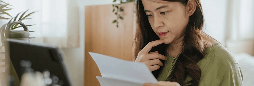 Eine Frau mit langen dunklen Haaren und grünem Oberteil sitzt an einem Tisch, hält ein Dokument in der Hand und stützt nachdenklich ihr Kinn.