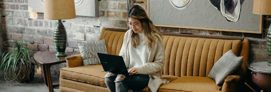 Eine Frau mit heller Strickjacke und Brille sitzt auf einem braunen Vintage-Sofa mit einem Laptop auf dem Schoß. Der Raum ist stilvoll mit Kunstwerken und Lampen dekoriert.