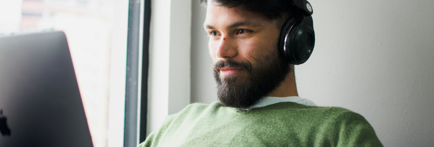 Ein junger Mann mit Bart und grünem Pullover sitzt am Fenster mit einem Laptop und trägt schwarze Over-Ear-Kopfhörer.