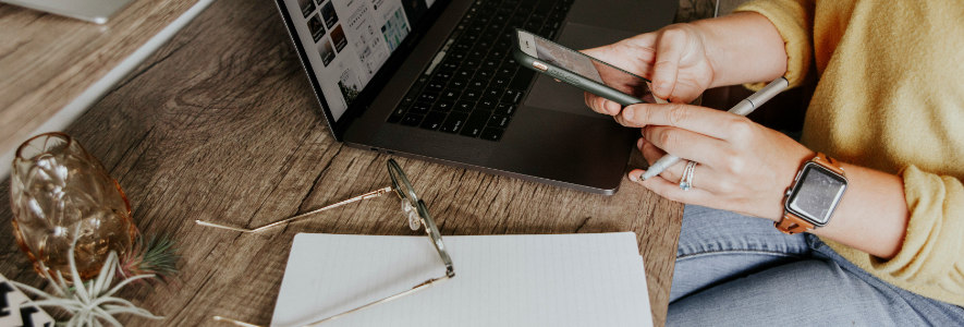 Eine Frau in gelbem Pullover sitzt an einem Holztisch mit Laptop, Smartphone und Notizblock. Sie tippt am Handy, während eine Brille und ein Glas daneben liegen.