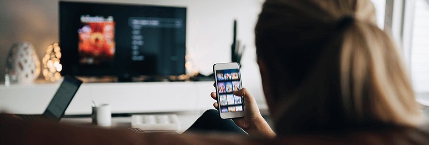 Eine Frau sitzt auf der Couch mit Smartphone in der Hand, während im Hintergrund der Fernseher mit einer Streaming-App geöffnet ist.