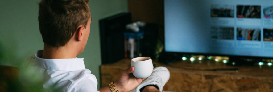 Ein junger Mann in weißem Hemd sitzt entspannt mit einer Tasse in der Hand vor dem Fernseher. Die Szene ist gemütlich beleuchtet.