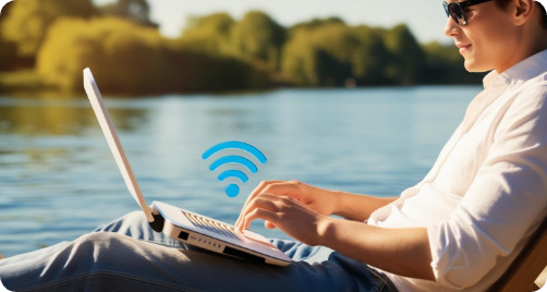 Mann mit Sonnenbrille arbeitet entspannt am Laptop am Seeufer mit WLAN-Symbol.